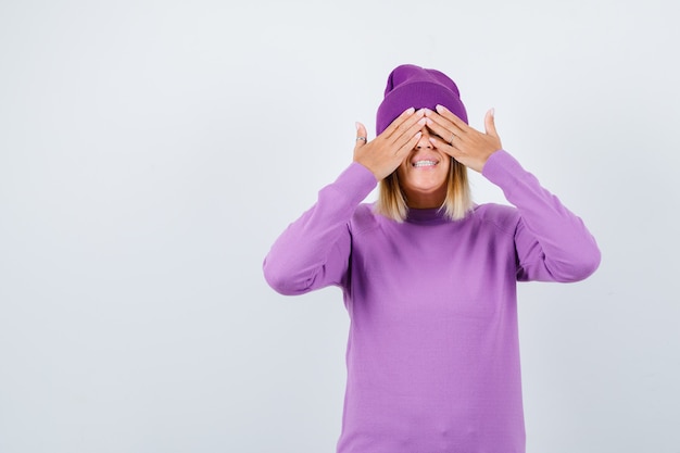 Señorita en suéter morado, gorro cubriendo los ojos con las manos y mirando alegre, vista frontal.