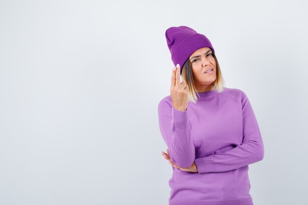 Señorita en suéter morado, gorro mostrando gesto de pistola y mirando confiado, vista frontal.