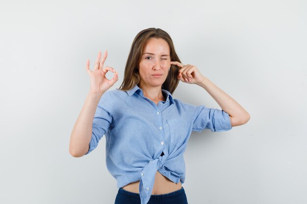 Señorita tapando la oreja, mostrando el signo de ok, guiñando el ojo en camisa azul