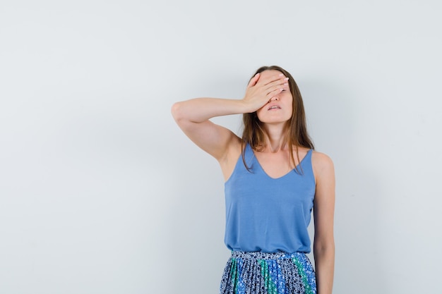 Señorita tapándose los ojos con la mano en blusa azul, falda y aspecto fatigado. vista frontal.