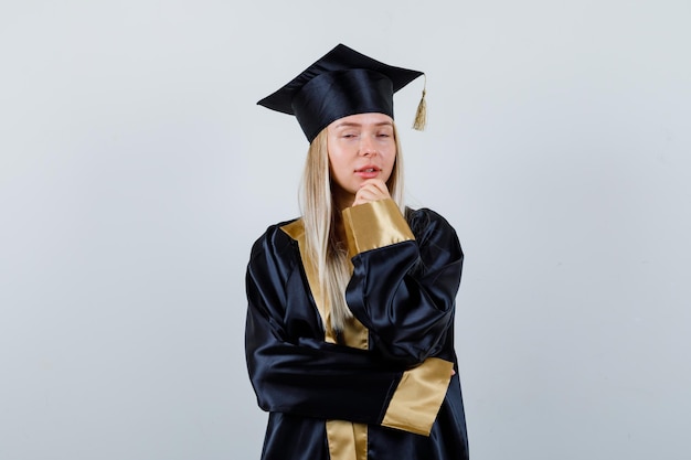 Señorita en traje académico de pie en pose de pensamiento y mirando pensativo