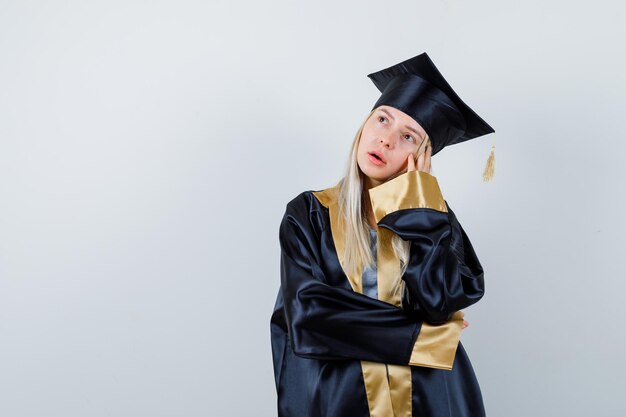 Señorita en traje académico de pie en pose de pensamiento y mirando pensativo