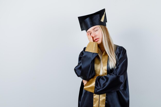 Señorita en traje académico sosteniendo la mano en la mejilla y con aspecto cansado