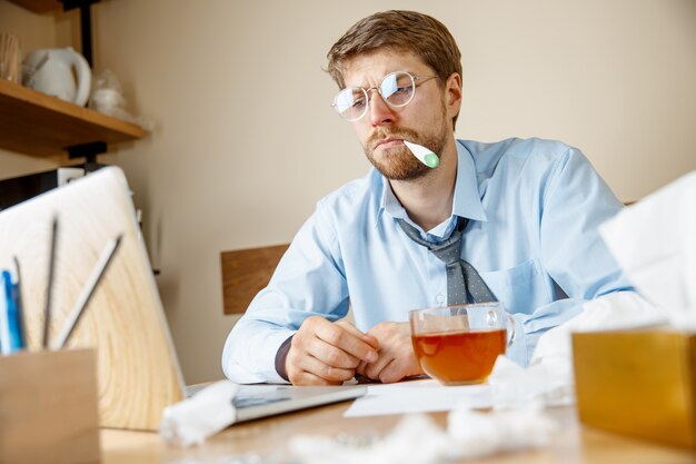 Sensación de malestar y cansancio. El hombre con una taza de té caliente que trabaja en la oficina, el empresario se resfrió, la gripe estacional. La influenza pandémica, la prevención de enfermedades, el aire acondicionado en la oficina causan enfermedades