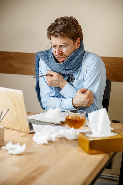 Sensación de malestar y cansancio. El hombre con una taza de té caliente que trabaja en la oficina, el empresario se resfrió, la gripe estacional. La influenza pandémica, la prevención de enfermedades, el aire acondicionado en la oficina causan enfermedades