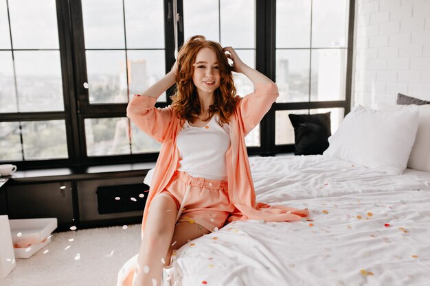 Sensual chica blanca en pijama sentada en la cama. Riendo a maravillosa dama posando en el dormitorio con una sonrisa feliz.