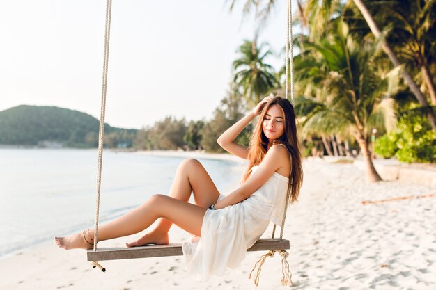 Sensual chica tierna sentada en un columpio con un vestido blanco. La niña tiene los ojos cerrados. Tiene el pelo largo y oscuro. Ella tiene brazaletes en su brazo y pierna. El columpio está en la playa con palmeras verdes.