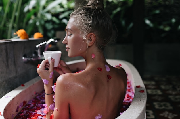 Sensual mujer blanca con cabello rubio bebiendo té en el baño. Increíble dama bronceada haciendo spa con los ojos cerrados y disfrutando de un café.