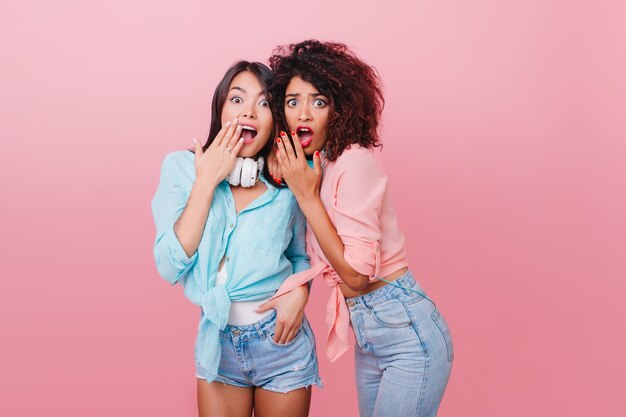 Sensual mujer caucásica con pelo corto negro posando con expresión de cara de sorpresa. Sorprendida mulata de pelo oscuro abrazando a una amiga latina.