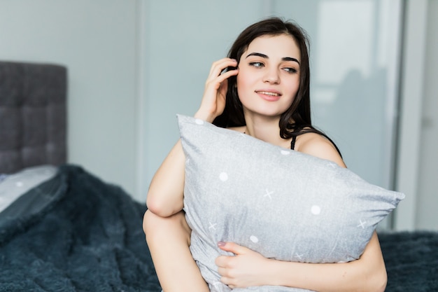 Sensual mujer joven sonriente sentada en la cama y abrazando la almohada blanca