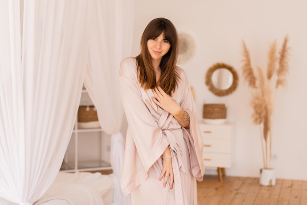 Sensual mujer morena posando en kimono de seda beige en elegante dormitorio ligero en estilo bohoh.