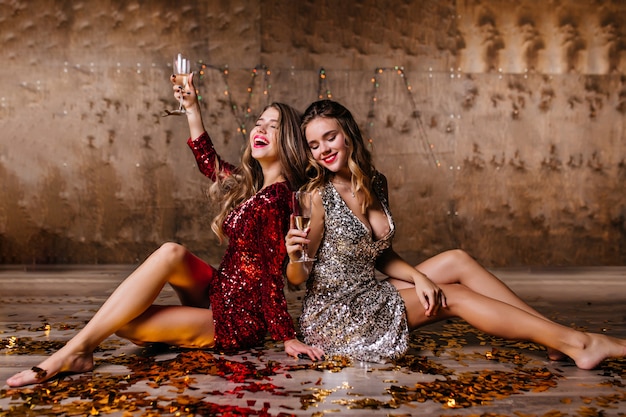 Sensual mujer rubia en vestido brillante bebiendo champán en el suelo