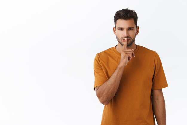 Sensual y soñador apuesto hombre barbudo en camiseta marrón callando y entrecerrando los ojos apasionadamente guardando secreto pidiendo silencio callando romántica y misteriosamente de pie fondo blanco