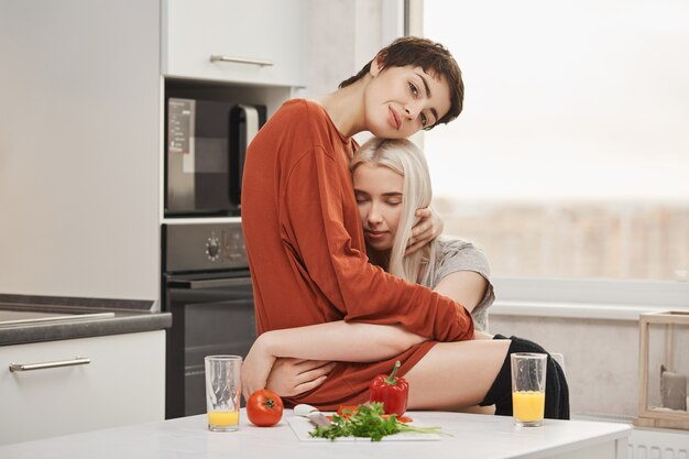 Sensual y tierna joven pareja de lesbianas enamoradas, abrazados en la cocina mientras desayunaban. La mujer está feliz por cada día que pasó con su novia, queriendo abrazarla y estar con ella para siempre.