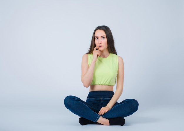 Sentada en el suelo, la chica está pensando sosteniendo el dedo índice en los labios sobre fondo blanco.