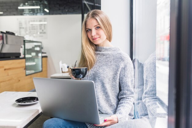 Sentado en café mujer usando laptop y sosteniendo la taza