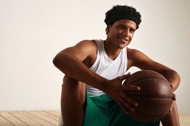 Sentado sonriente amigable jugador de baloncesto afroamericano con un afro vistiendo uniforme blanco y verde sosteniendo una pelota de cuero marrón