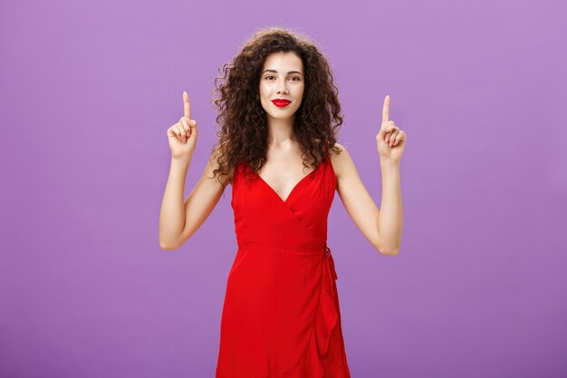 Sentirse en la cima después de hacer un maquillaje elegante. Linda y tierna mujer europea glamorosa en vestido de noche rojo y lápiz labial con cabello rizado oscuro sonriendo suavemente y apuntando hacia arriba con las manos levantadas.