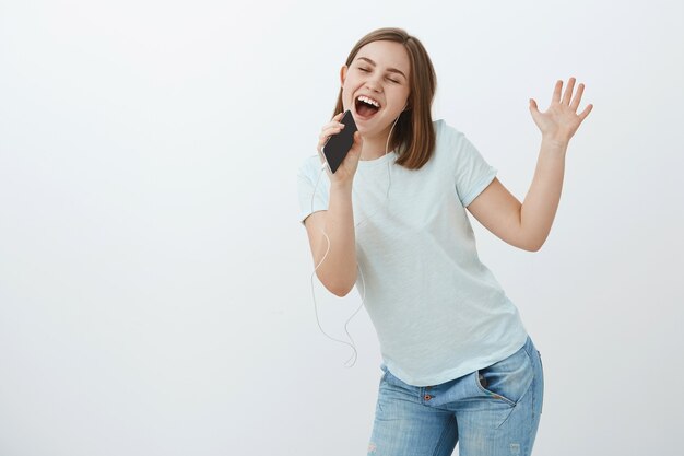 Sentirse como cantante popular en el escenario. Mujer divertida y energizada en camiseta bailando inclinando el cuerpo y agitando la cabeza escuchando música en auriculares con los ojos cerrados cantando en el teléfono inteligente como en el micrófono