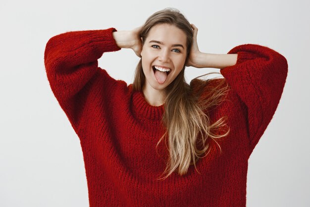 Sentirse independiente y sin preocupaciones. Retrato de mujer caucásica infantil complacida en elegante atuendo, tomados de la mano en la cabeza y mostrando la lengua, sonriendo ampliamente, bromeando con amigos