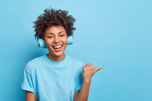 Será mejor que sigas este camino. Sonriente niña afroamericana de piel oscura apunta con el pulgar hacia afuera sugiere un lugar para visitar usa audífonos inalámbricos vestidos casualmente disfruta escuchando música a través de audífonos