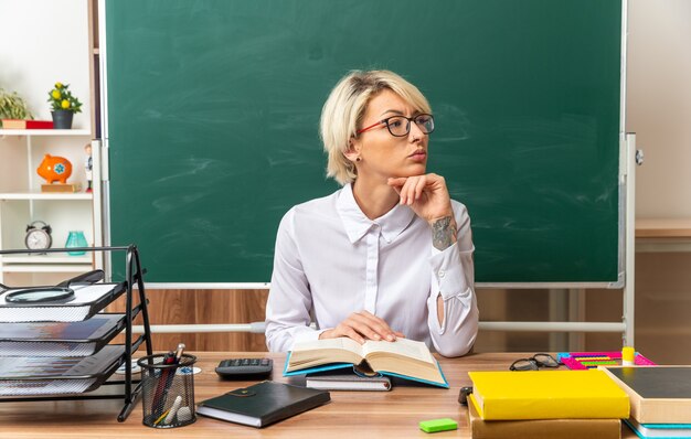 seria joven rubia maestra con gafas sentado en el escritorio con útiles escolares en el aula manteniendo la mano debajo de la barbilla y en el libro abierto mirando de lado