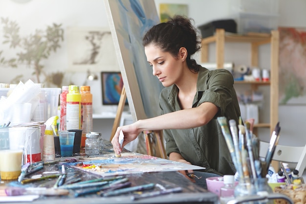 Foto gratuita seria morena joven y bella mujer sentada en un estudio de arte, tomando pinturas de colores del tubo mientras crea una gran obra maestra en el caballete, se preocupa por su trabajo y tiene una buena imaginación