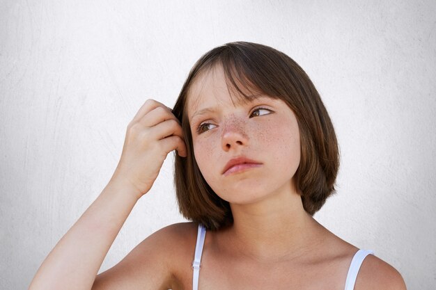 Seria y pequeña niña pecosa, manteniendo su mano sobre el cabello, teniendo una expresión pensativa, mirando a un lado. Hermosa chica posando contra la pared blanca. Concepto de belleza, infancia, expresión facial