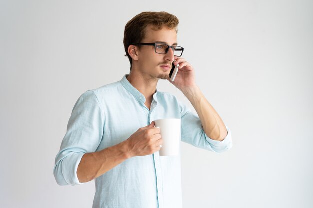 Serio apuesto joven guapo con taza hablando por teléfono.