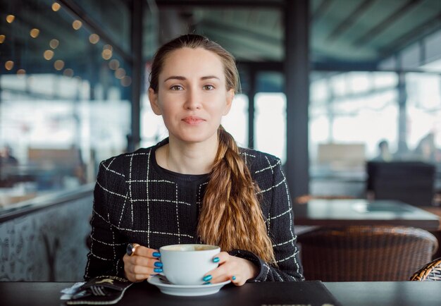 En serio, la chica bloguera está mirando a la cámara sosteniendo una taza de café en la cafetería