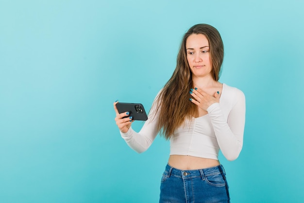 En serio, la chica está mirando la pantalla del teléfono inteligente sosteniendo la mano en el pecho sobre fondo azul.