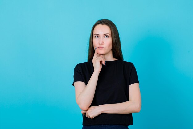 En serio, la chica está pensando sosteniendo el dedo índice en la mejilla sobre fondo azul.