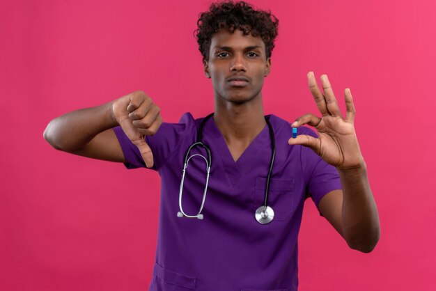 Un serio joven apuesto médico de piel oscura con cabello rizado vistiendo uniforme violeta con estetoscopio mostrando los pulgares hacia abajo mientras sostiene la píldora