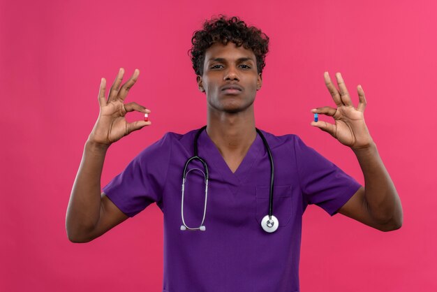 Un serio joven apuesto médico de piel oscura con cabello rizado vistiendo uniforme violeta con estetoscopio sosteniendo pastillas