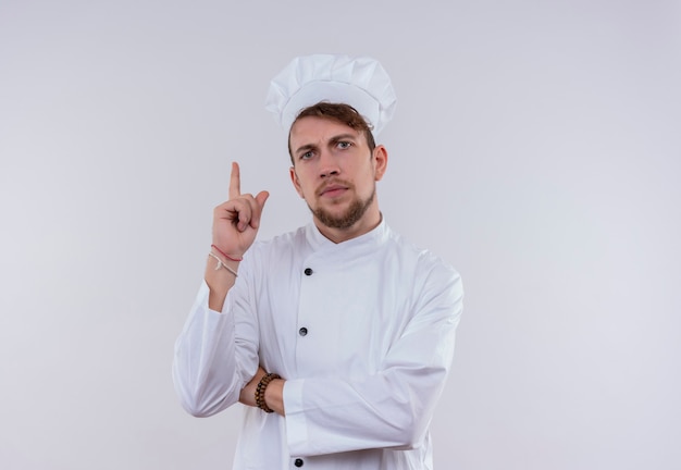 Un serio joven chef barbudo con uniforme blanco apuntando hacia arriba con las manos cruzadas mientras mira en una pared blanca