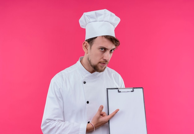 Foto gratuita un serio joven chef barbudo con uniforme blanco apuntando a la carpeta en blanco mientras mira en una pared rosa