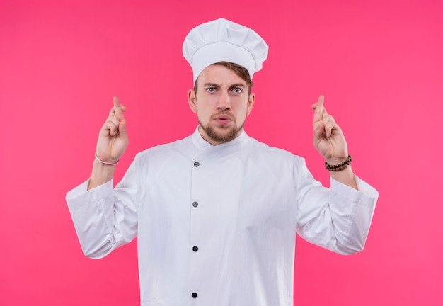 Un serio joven chef barbudo en uniforme blanco cruzando el dedo medio sobre el dedo índice en una pared rosa