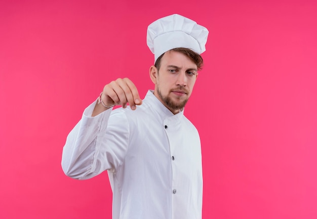 Un serio joven chef barbudo en uniforme blanco vistiendo gorro de cocinero espolvoreando especias en el plato sobre una pared rosa