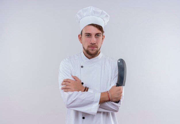 Un serio joven chef barbudo vestido con uniforme de cocina blanco y sombrero sosteniendo cuchillo de carnicero mientras mira en una pared blanca