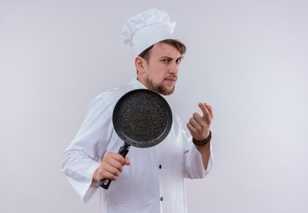 Un serio joven chef barbudo vestido con uniforme de cocina blanco y sombrero sosteniendo una sartén como un bate de béisbol en una pared blanca