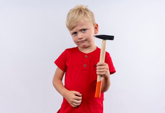 Un serio niño rubio lindo en camiseta roja sosteniendo un martillo mientras mira en una pared blanca