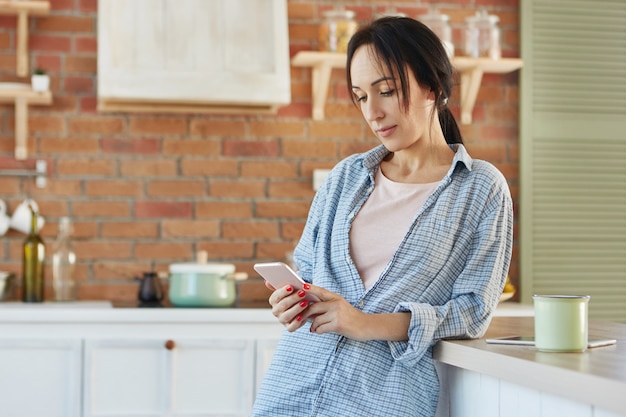 Serios mensajes de mujer morena en línea, usa conexión gratuita a Internet, usa camisa informal,