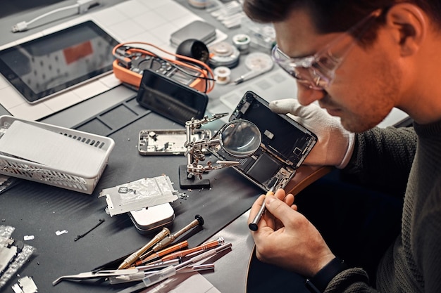 Foto gratuita serviceman usa lupa y destornillador para reparar el teléfono inteligente dañado en el taller electrónico.