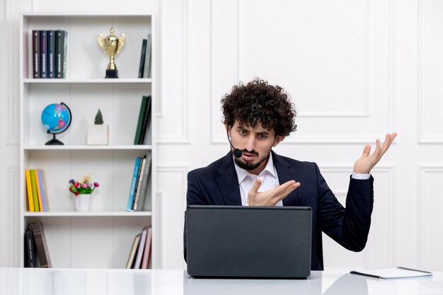 Servicio al cliente apuesto hombre rizado en traje de oficina con computadora y auriculares molesto