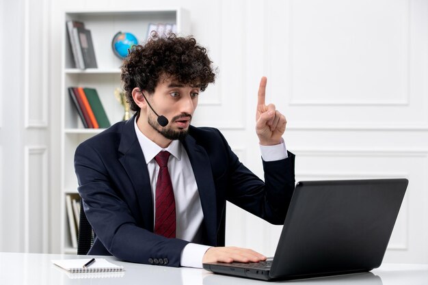 Servicio al cliente apuesto joven en traje de oficina con computadora portátil y auriculares apuntando hacia arriba