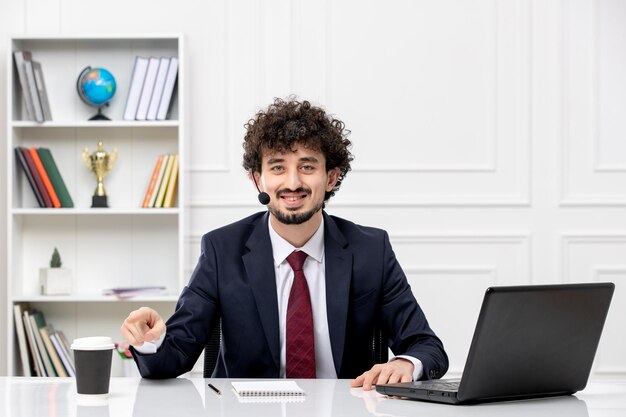 Servicio al cliente apuesto joven en traje de oficina con computadora portátil y auriculares apuntando a la taza de café