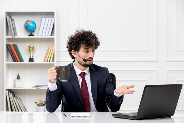 Servicio al cliente apuesto joven en traje de oficina con computadora portátil y auriculares confundidos con taza