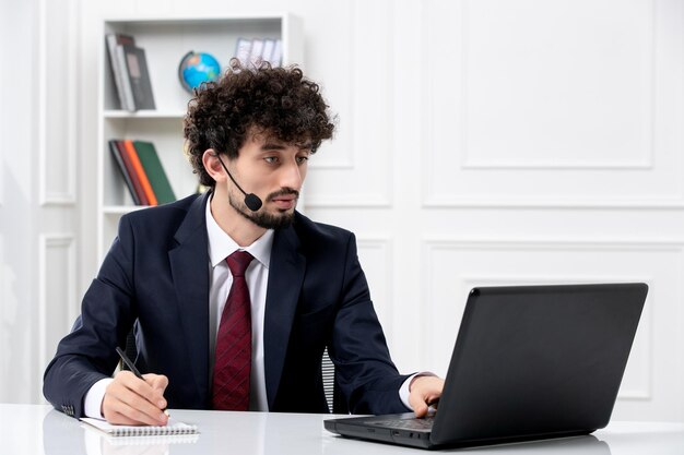 Servicio al cliente apuesto joven en traje de oficina con computadora portátil y auriculares escribiendo notas