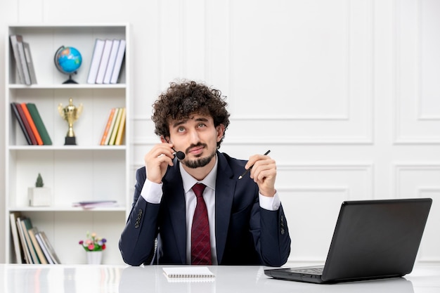 Servicio al cliente apuesto joven en traje de oficina con computadora portátil y auriculares mirando hacia arriba y pensando