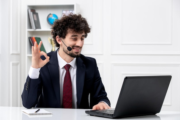 Servicio al cliente apuesto joven en traje de oficina con computadora portátil y auriculares que muestran un gesto correcto